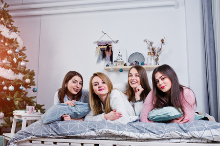 Four cute friends girls wear on warm sweaters and black pants on bed at new year decorated room on studio, play with pillows.の写真素材