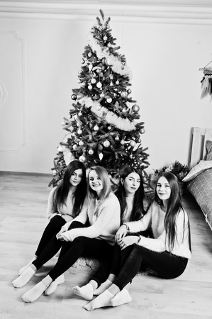 Four cute friends girls wear on warm sweaters, black pants against new year tree with christmas decoration at white room.の写真素材