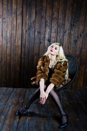 Elegante blonde girl wear on fur coat sitting on chair at studio against wooden background.の写真素材
