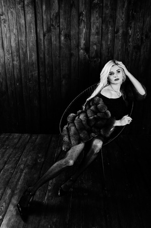 Elegante blonde girl wear on fur coat sitting on chair at studio against wooden background.の写真素材