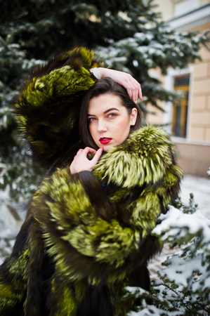 Brunette girl in green fur coat at winter day against snowy pine tree.の写真素材