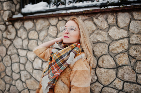 Portraiy of blonde girl in glasses, red fur coat and scarf at winter day.の写真素材