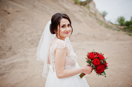 Portrait of a bride holding a wedding bouquet of red flowers and posing alone in sand quarry.の写真素材