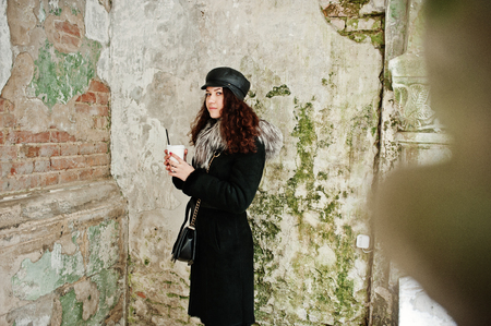 Curly mexican girl in leather cap and plastic cup of coffee at hand walking at streets of city.の写真素材
