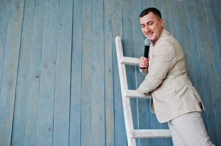 Handsome man in beige suit and pink shirt with microphone against ladder background on studio. Funny face of toastmaster and showman.の写真素材