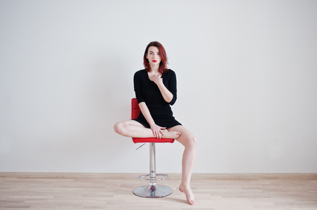 Red haired girl on black dress tunic sitting on red chair against white wall at empty room.の写真素材