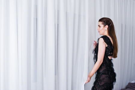 Brunette girl in black evening dress against white room with bed.の写真素材