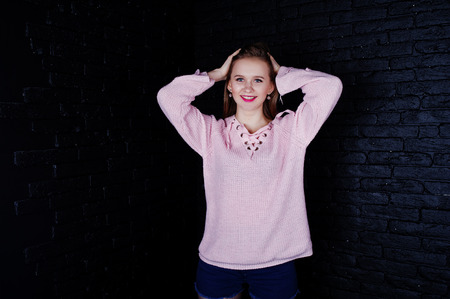 Studio shot of brunette girl at pink sweater with jeans shorts against black brick wall.