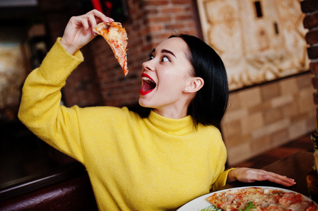 Funny brunette girl in yellow sweater eating pizza at restaurant. の写真素材