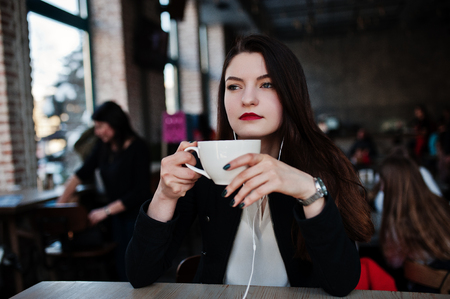 Brunette girl sitting on cafe with cup of cappuccino, listening music on headphones.の写真素材