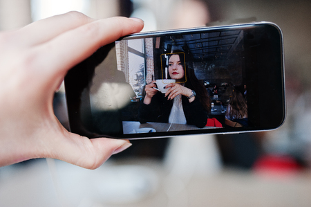 Close up photo of screen mobile phones with brunette girl sitting on cafe with cup of cappuccino, listening music on headphones.の写真素材