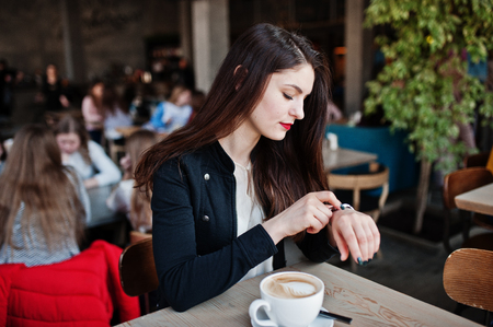 Brunette girl sitting on cafe with cup of cappuccino and looking at her watches on hand.の写真素材