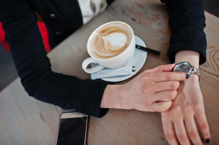 Close up photo of girl sitting on cafe with cup of cappuccino and looking at her watches on hand.の写真素材