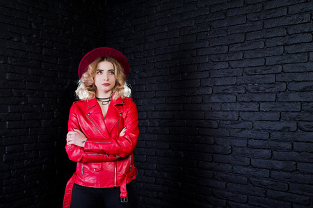 Studio portrait of blonde girl in red hat and leather jacket against brick wall.の写真素材