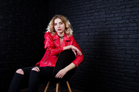 Studio portrait of blonde girl in red leather jacket posed on chair against brick wall.の写真素材