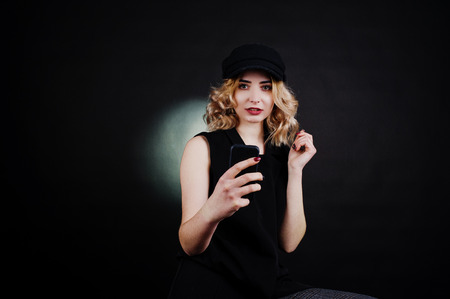 Studio portrait of blonde girl in black wear and cap with mobile phone at hand against dark...