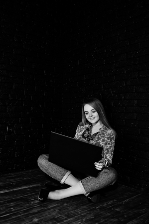 Stylish blonde girl in jacket and jeans with laptop against brick black wall at studio.の写真素材