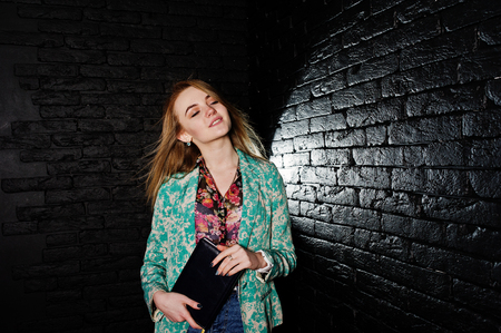 Stylish blonde girl in jacket and jeans with notebook diary at hands against brick black wall at studio.の写真素材