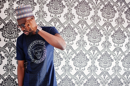 Rich african man in stylish traditional clothes and hat in glasses posed at studio against vintage...