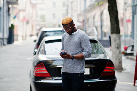 Stylish african american boy on gray sweater and glasses posed at street against black business car and speaking on phone. Fashionable black guy.の写真素材