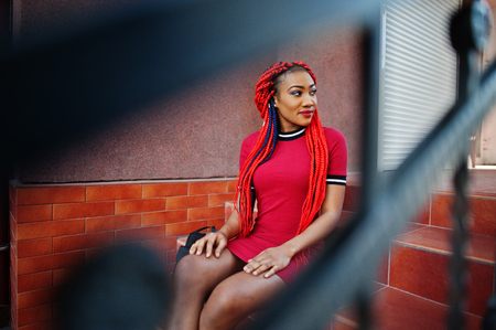 Cute and slim african american girl in red dress with dreadlocks and backpack posed outdoor, sitting on stairs. Stylish black model.の写真素材