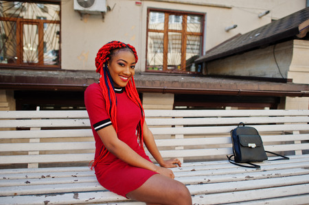 Cute and slim african american girl in red dress with dreadlocks and backpack posed outdoor on street, sitting on bench. Stylish black model.の写真素材