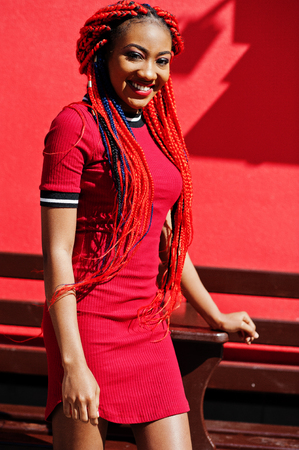 Cute and slim african american girl in red dress with dreadlocks posed outdoor against red wall. Stylish black model.の写真素材