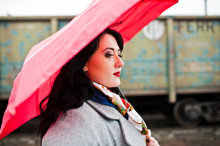 Brunette girl in gray coat with red umbrella in railway station.の写真素材