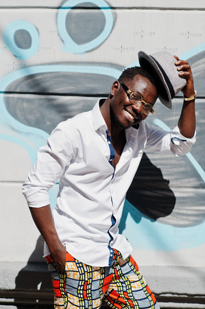 Stylish african american man in white shirt and colored pants with hat and glasses posed outdoor against graffiti wall. Black fashionable model boy.の写真素材