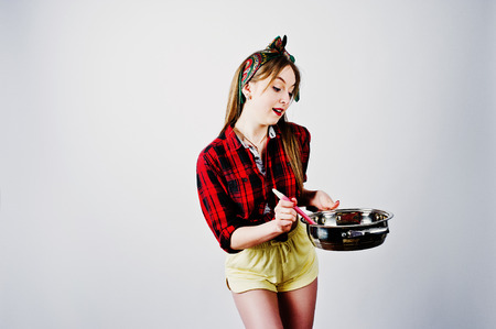 Young funny housewife in checkered shirt and yellow shorts pin up style with frying pan isolated on white background.の写真素材