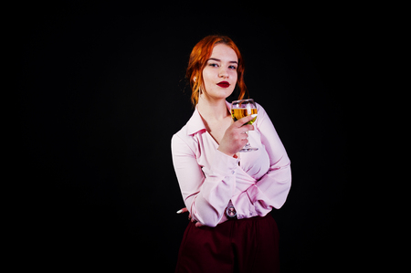 Gorgeous red haired girl in pink blouse and red skirt with glass of wine at hand isolated on black.の写真素材