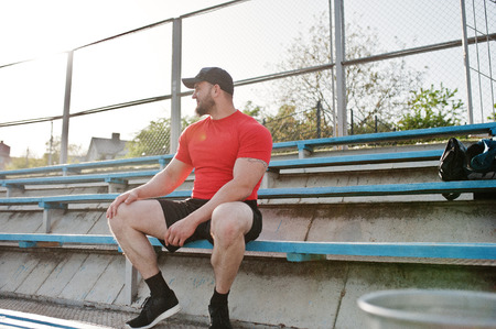 Young brutal bearded muscular man wear on red shirt, shorts and cap at stadium.の写真素材