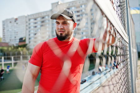 Young brutal bearded muscular man wear on red shirt, shorts and cap at stadium.の写真素材