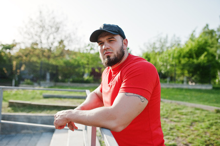 Young brutal bearded muscular man wear on red shirt, shorts and cap at stadium.
