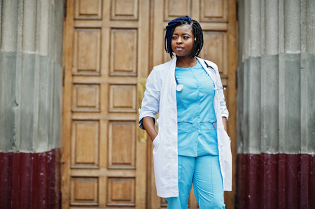 Stylish african american doctor with stethoscope and lab coat posed against door of hospital.の写真素材