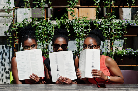 Three stylish african american womans posed at sunny summer day outdoor, sitting on table of restaurant with menu at hands.の写真素材