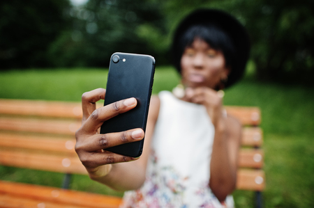 Amazing african american model woman in green pants and black hat posed on bench at park and making selfie on mobile phone.の写真素材