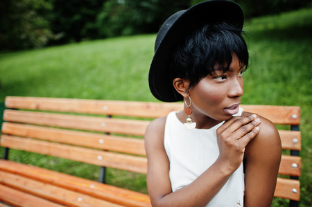 Amazing african american model woman in green pants and black hat posed on bench at park.