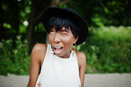 Amazing african american model woman in green pants and black hat posed at park, wink.