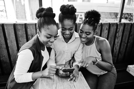 Three african american girls sitting on the table of caffe and looking on mobile phone.の写真素材