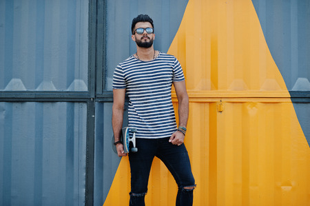 Handsome tall arabian beard man model at stripped shirt posed outdoor. Fashionable arab guy at sunglasses with skateboard against yellow painted wall.の写真素材