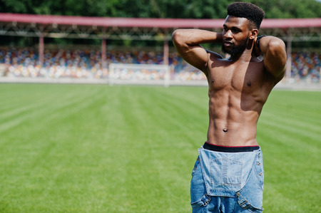 Handsome sexy african american muscular bare torso man at jeans overalls posed at green grass of stadium football field. Fashionable black man portrait.の写真素材