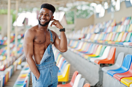 Handsome sexy african american bare torso man at jeans overalls posed on colored chairs at stadium. Fashionable black man portrait.の写真素材