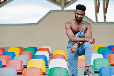 Handsome sexy african american bare torso man at jeans overalls posed on colored chairs at stadium. Fashionable black man portrait.の写真素材