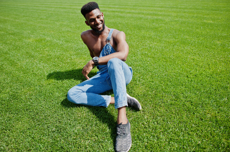 Handsome sexy african american bare torso man at jeans overalls sitting at green grass of stadium football field. Fashionable black man portrait.の写真素材