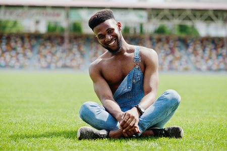 Handsome sexy african american bare torso man at jeans overalls sitting at green grass of stadium football field. Fashionable black man portrait.の写真素材