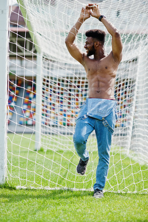 Handsome sexy african american muscular bare torso man at jeans overalls posed at green grass agasinst football gates of stadium field. Fashionable black man portrait.の写真素材