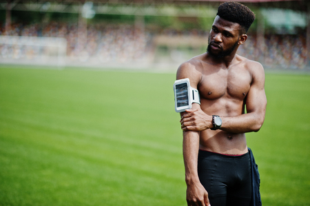 African american male athlete sexy sport bare torso man with running sports arm case for mobile phone, posed at green grass of football stadium.の写真素材