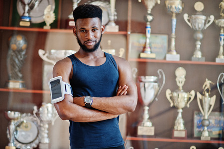 African american male athlete sexy sport man with running sports arm case for mobile phone, posed against many golden cups.の写真素材