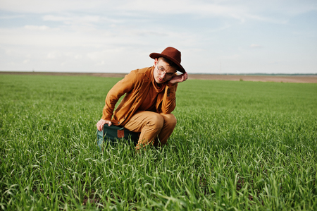 Stylish man in glasses, brown jacket and hat with bag posed on green field.の写真素材